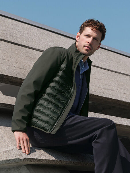 Beige sneakers with serrated soles worn with a high-necked down jacket and dark men's trousers resting on modern architecture | GEOX | GEOX